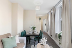 A dining area with a table set for four, green upholstered chairs, a hanging plant, and a view through large windows. - Stylish Brick Lane Apartment Sleeps 9 Shoreditch (Greater London)