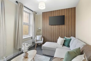 A modern living room with a sofa, a flat-screen TV mounted on a wooden panel wall, a chair, and a table with a vase of flowers. - Stylish Brick Lane Apartment Sleeps 9 Shoreditch (Greater London)