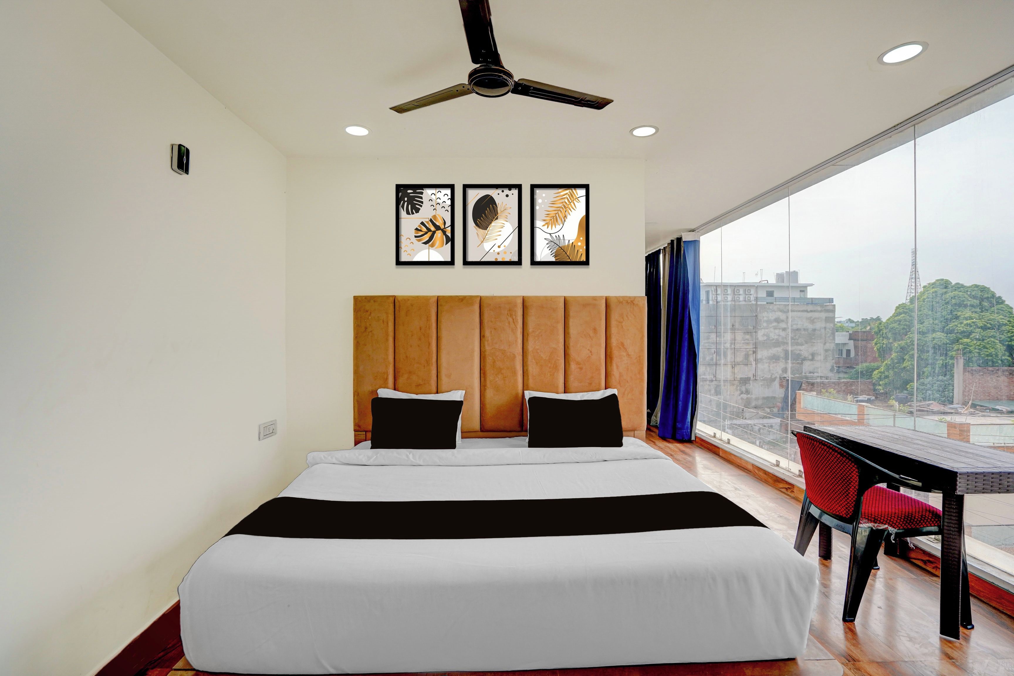 A hotel room with a bed, ceiling fan, artwork, and a view of the city.