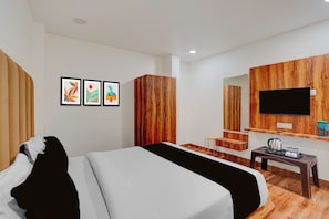 Habitación de hotel con una cama, un televisor montado en un panel de madera, una mesita con una tetera y un espejo sobre la mesita.