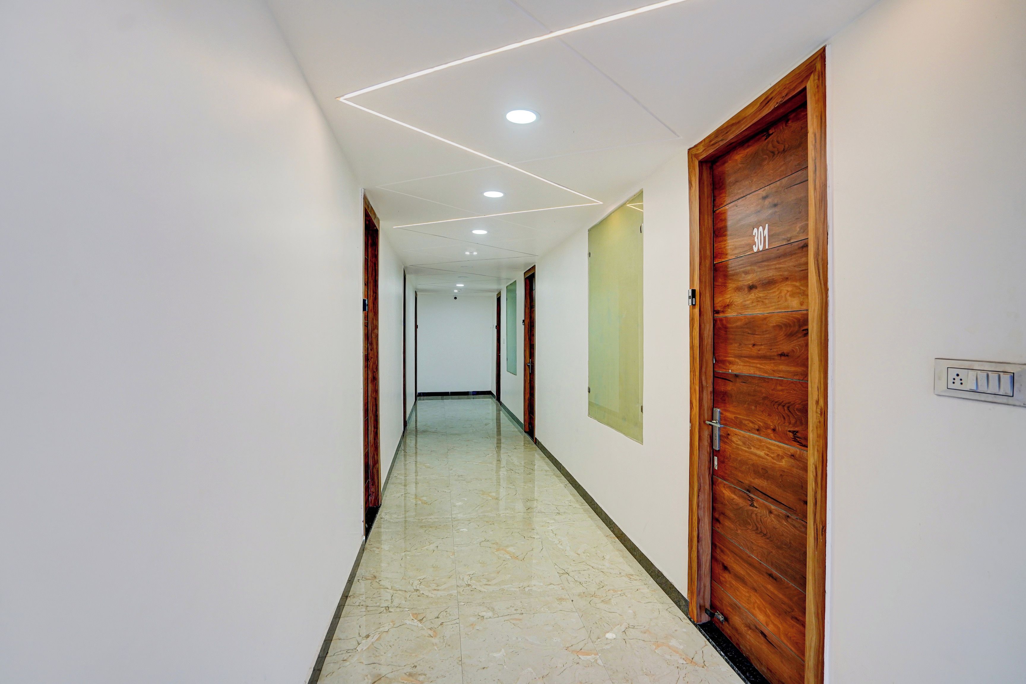 A corridor with a wooden door on the right, a light switch on the wall, and a ceiling with recessed lighting.