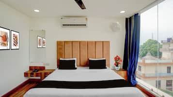 A hotel room with a large bed, a ceiling fan, an air conditioning unit, a bedside table with a phone, and a window with a view of buildings.