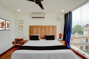 A hotel room with a large bed, a ceiling fan, an air conditioning unit, a bedside table with a phone, and a window with a view of buildings.