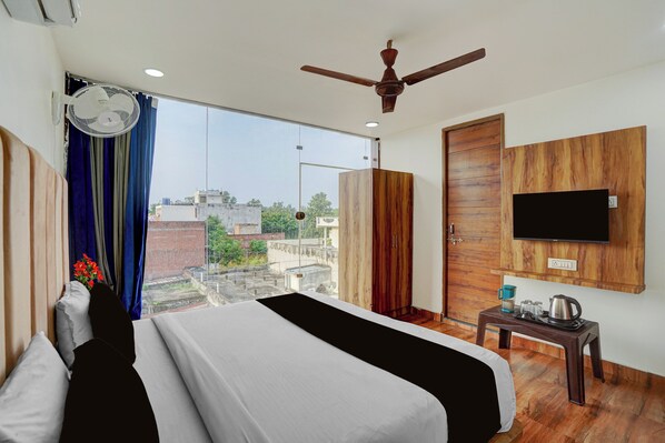 Habitación de hotel con cama, ventilador de techo, televisor montado en la pared y vista de la ciudad a través de amplias ventanas.