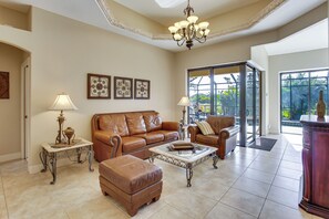 A spacious living room with a brown leather sofa, matching ottoman, and a glass coffee table. There are two side tables with lamps, and a chandelier hangs from the ceiling. The room has large windows with a view of greenery outside. - Lanai w/ Pool & Spa! Rotonda West Retreat (Rotonda West)