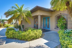 A residential building with a blue door, a small porch with a table and chairs, and a well-maintained garden with various plants and a palm tree. - Lanai w/ Pool & Spa! Rotonda West Retreat (Rotonda West)