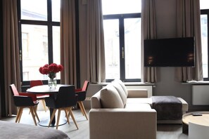 En moderne stue med sofa, spisebord med stole, fladskærms-tv på væggen og store vinduer med gardiner.