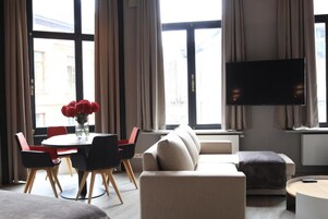 A modern living room with a sofa, a dining table with chairs, a flat-screen TV mounted on the wall, and large windows with curtains.