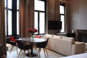A modern living room with a sofa, a dining table with chairs, a flat-screen TV mounted on the wall, and a fireplace.