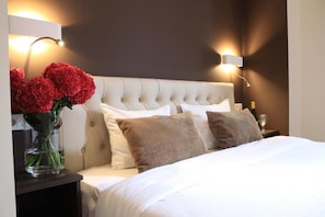 Una habitación de hotel con cabecera capitonada, una mesita de noche con un florero de flores rojas y dos lámparas de pared.