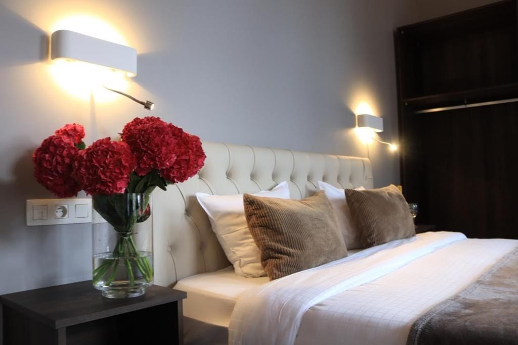 A bedroom with a bed, a nightstand, a vase with flowers, and wall-mounted lighting.