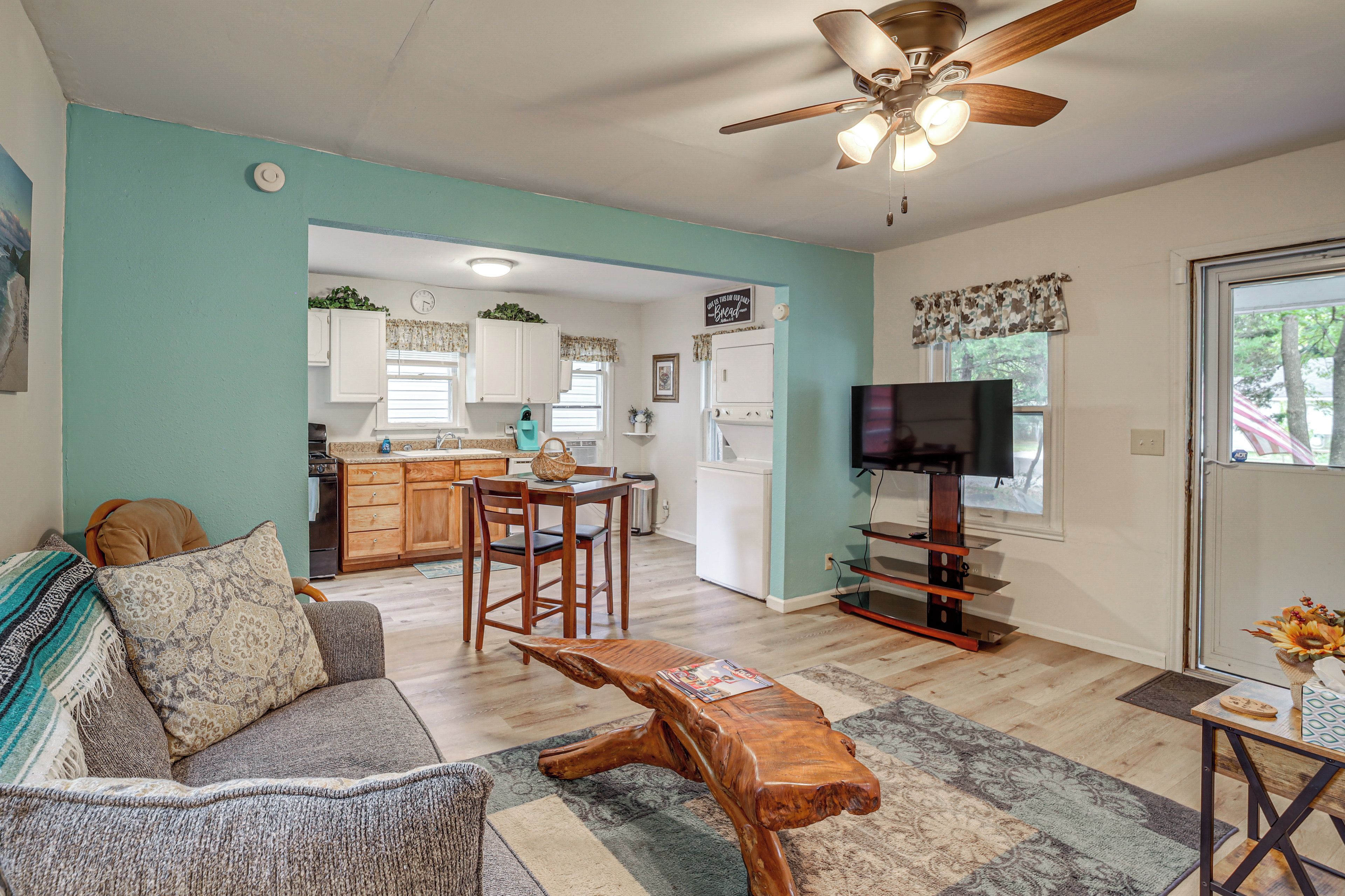 A living room with a sofa, coffee table, and a television. There is a kitchen area with cabinets and a refrigerator.