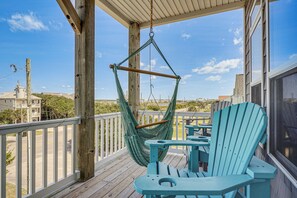 Eine Holzterrasse mit einer Hängematte, türkisfarbenen Adirondack-Stühlen und Blick auf das Meer und Gebäude.