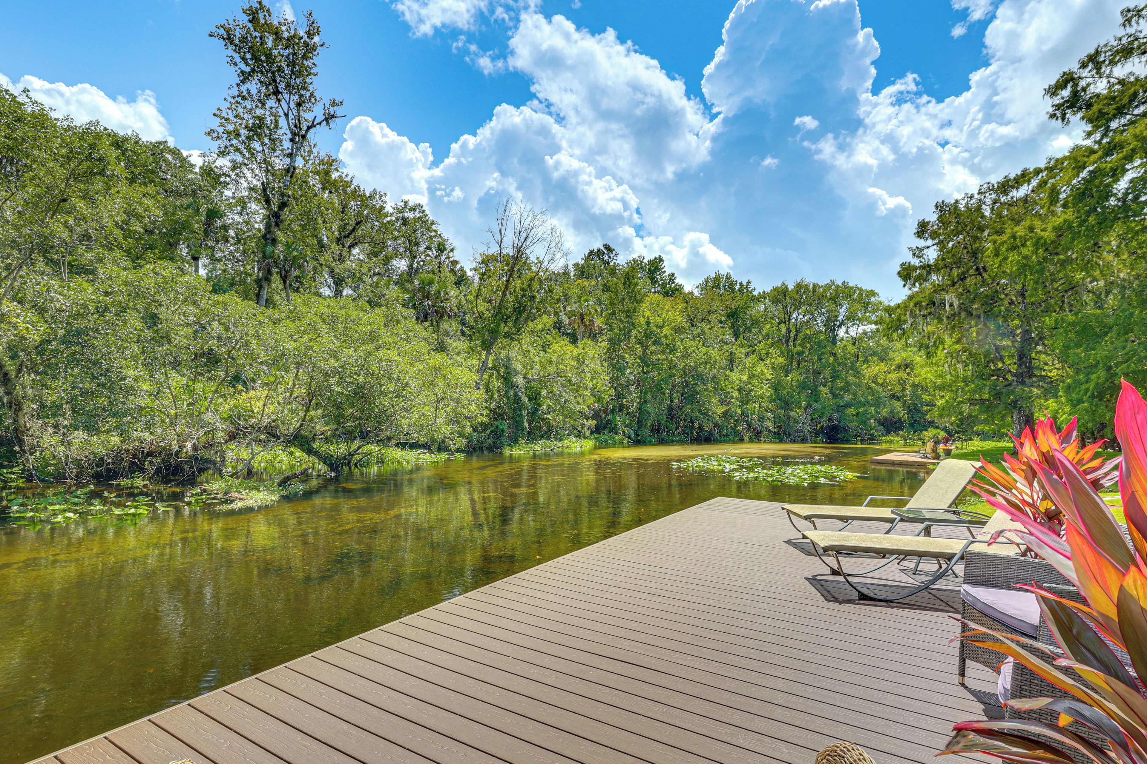 Een houten terras met lounge stoelen met uitzicht op een rustige rivier, omringd door weelderige begroeiing en bomen.