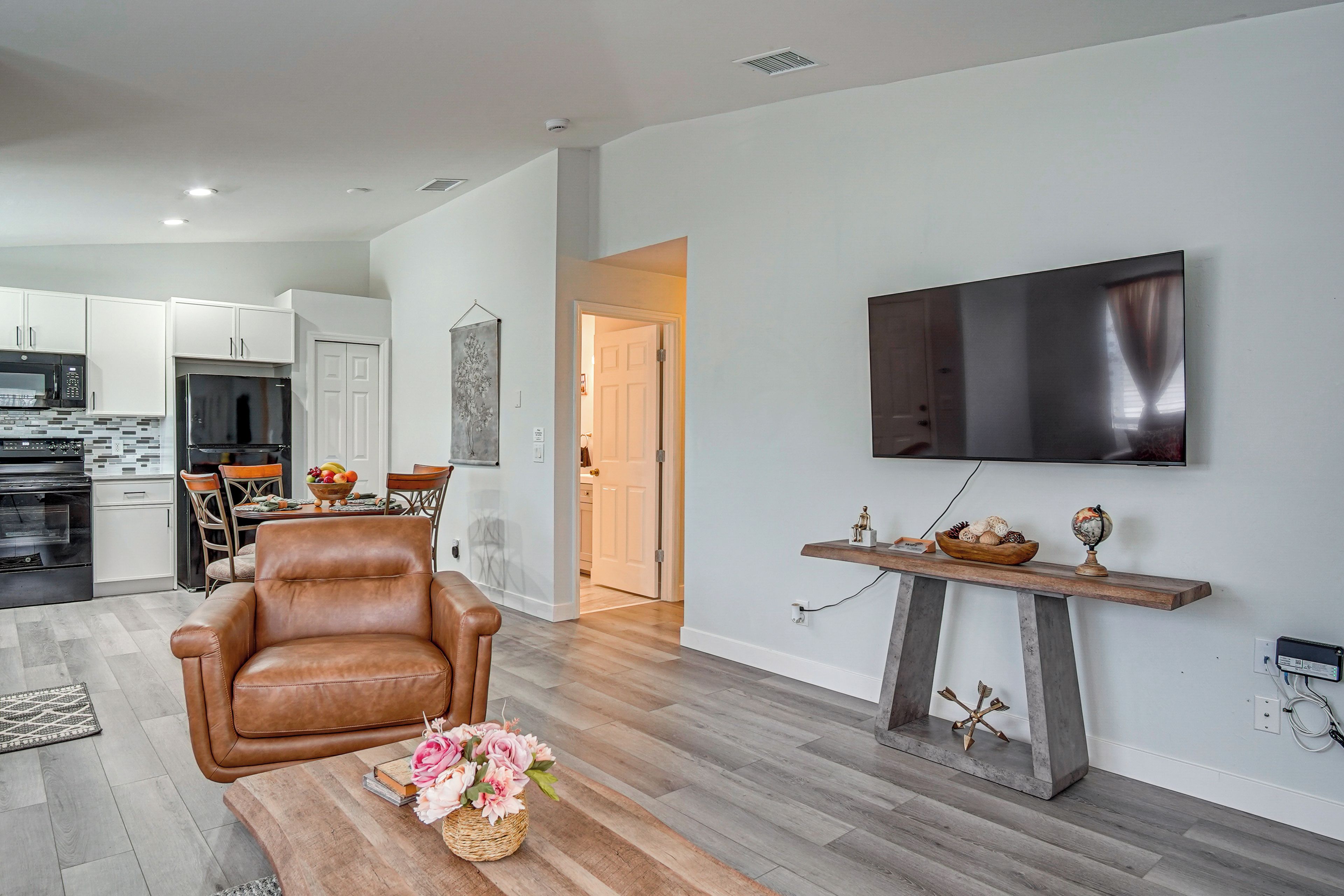 A modern living room with a leather armchair, a coffee table with a floral arrangement, a mounted flat-screen TV, and a wooden shelf with decorative items.