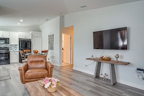 A modern living room with a leather armchair, a coffee table with a floral arrangement, a mounted flat-screen TV, and a wooden shelf with decorative items.