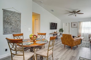 A dining area with a round table, chairs, and a fruit bowl. There is a television mounted on the wall and a ceiling fan.