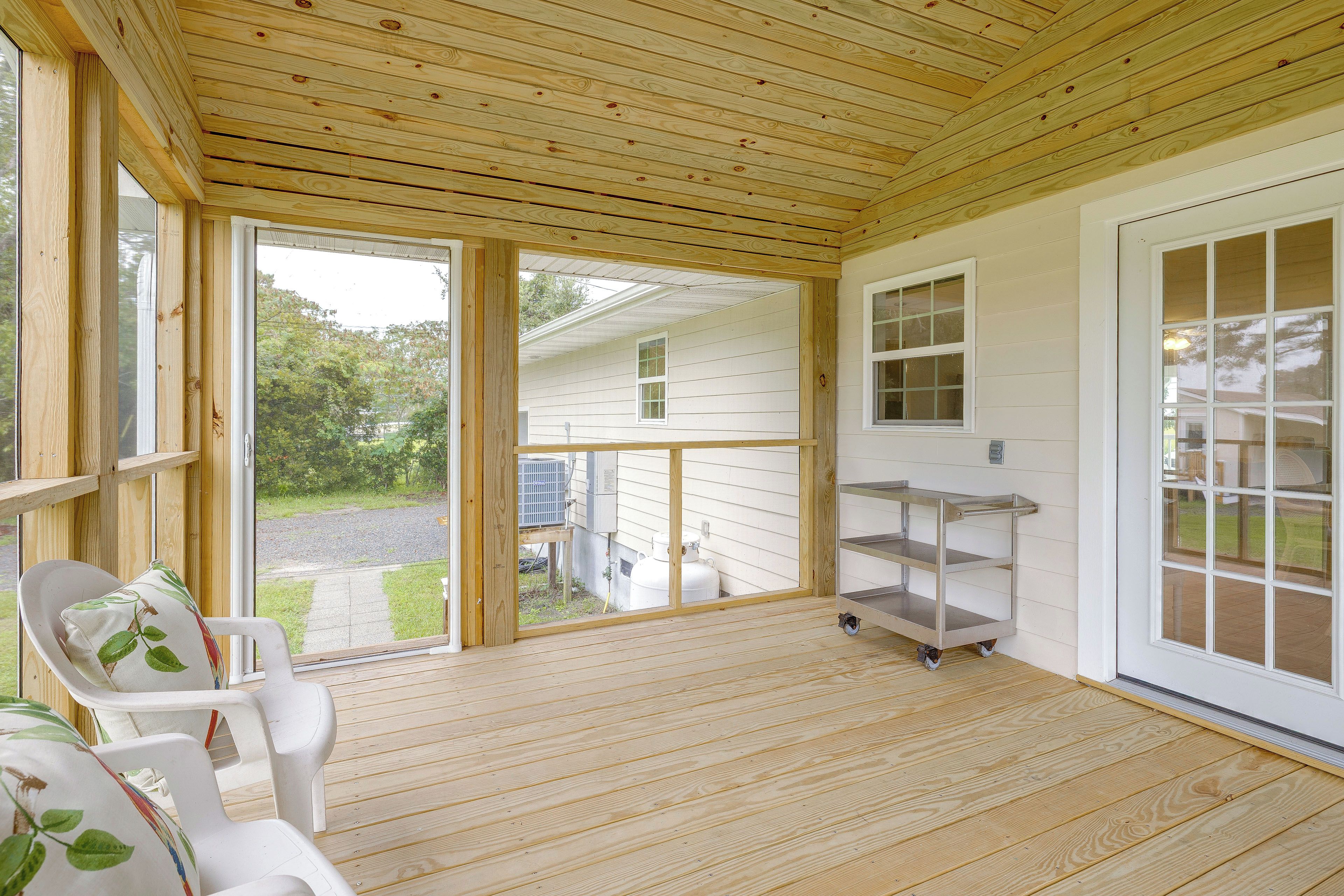 A covered porch with a wooden floor, a white chair with a leaf-patterned cushion, and a metal cart.