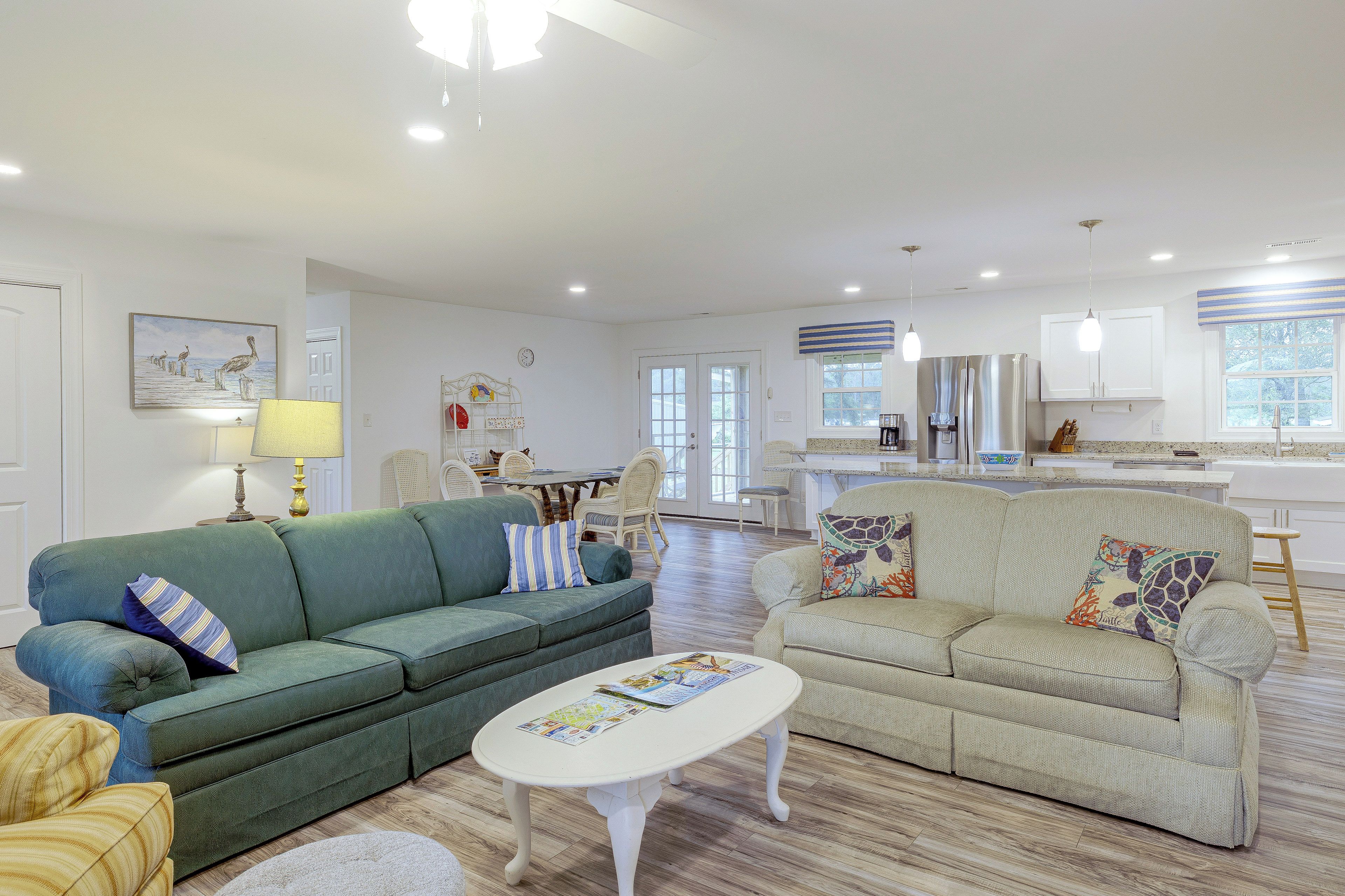 A spacious living room with a green sofa, a beige sofa, and a white coffee table. There is a dining area with a table and chairs in the background.