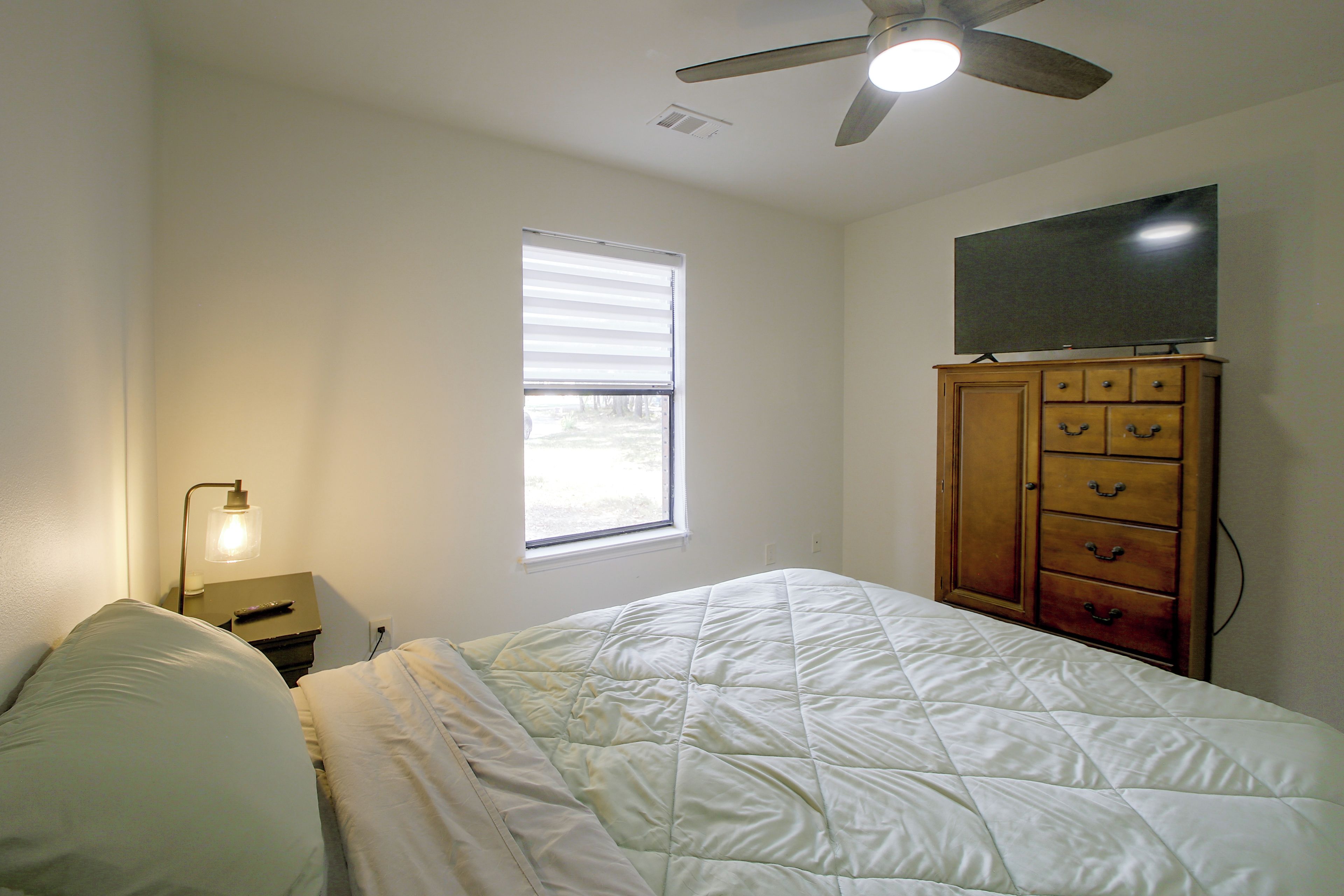 Un dormitorio con cama, ventilador de techo, mesita de noche con lámpara, cómoda y ventana.