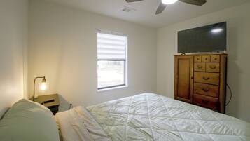 Un dormitorio con cama, ventilador de techo, mesita de noche con lámpara, cómoda y ventana.