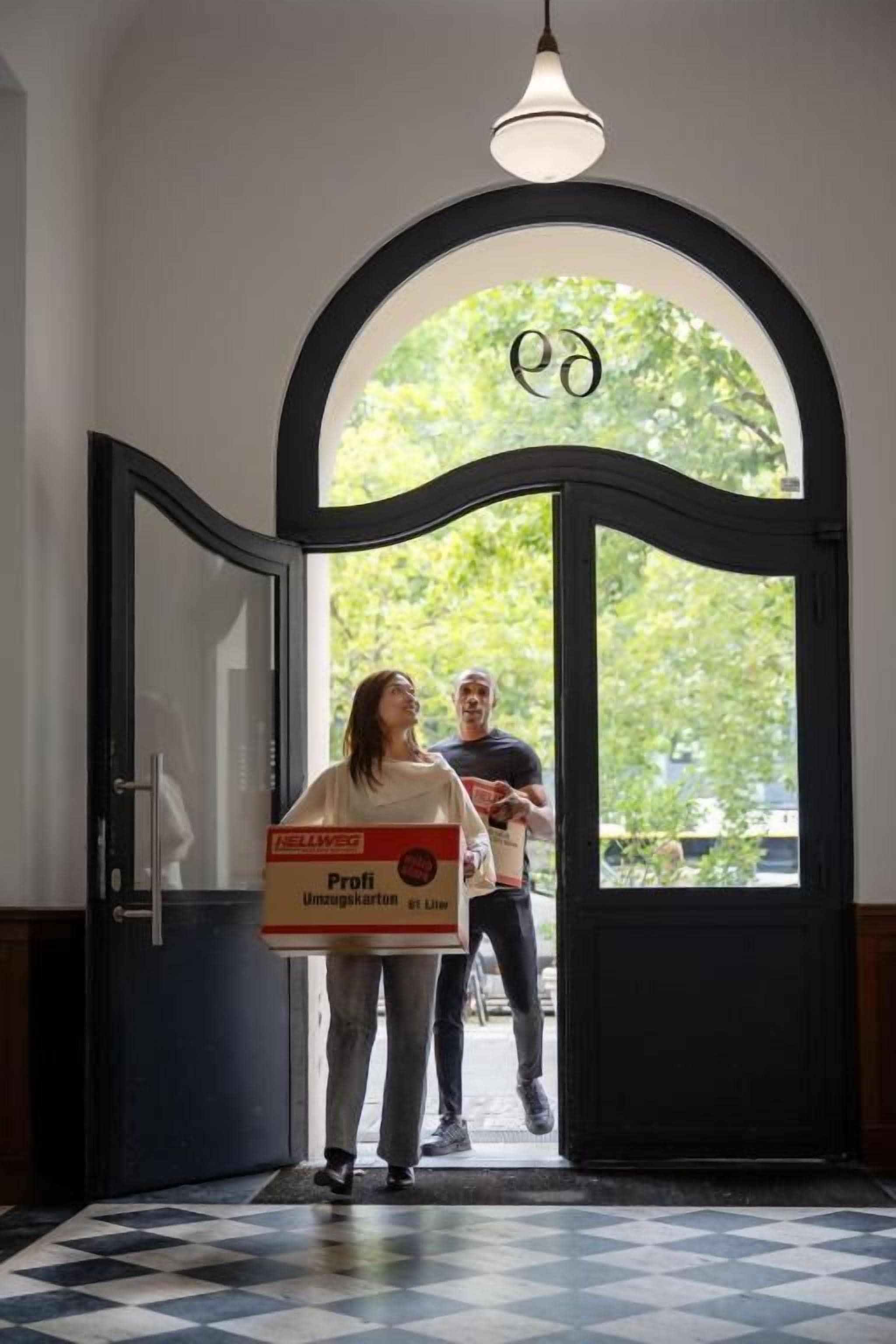 Una persona sosteniendo una caja con texto, de pie en un umbral con un arco circular y una lámpara colgante.