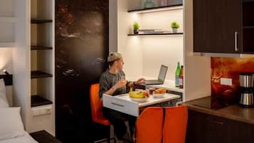 Ein kleines Zimmer mit einem Bett, einem Schreibtisch mit einem Laptop, einem Stuhl, einem kleinen Tisch mit Essen und einer Kaffeemaschine.