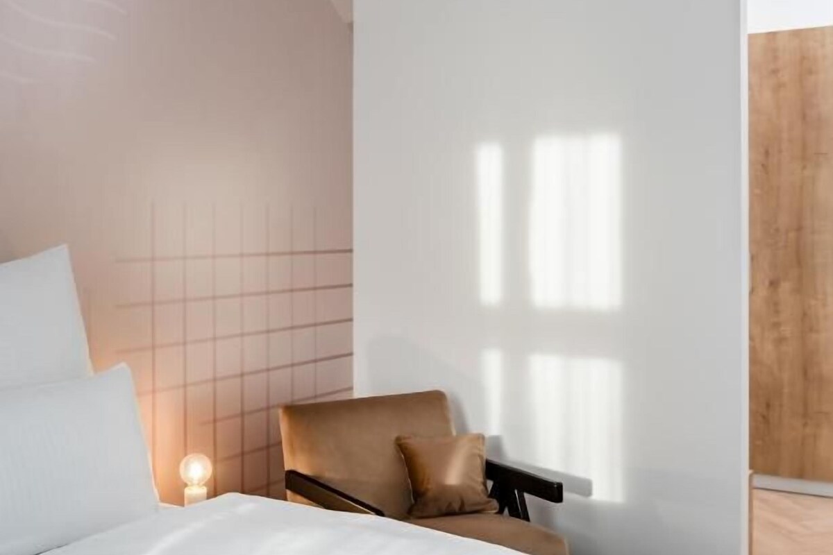 A modern hotel room with a bed, a chair, and a nightstand with a lamp.