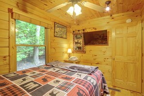 A wooden cabin room with a bed, a window, a ceiling fan, and a wall-mounted TV. - Pet-friendly Cozy Cabin Retreat Hot Tub & Trails (Cherry Log)