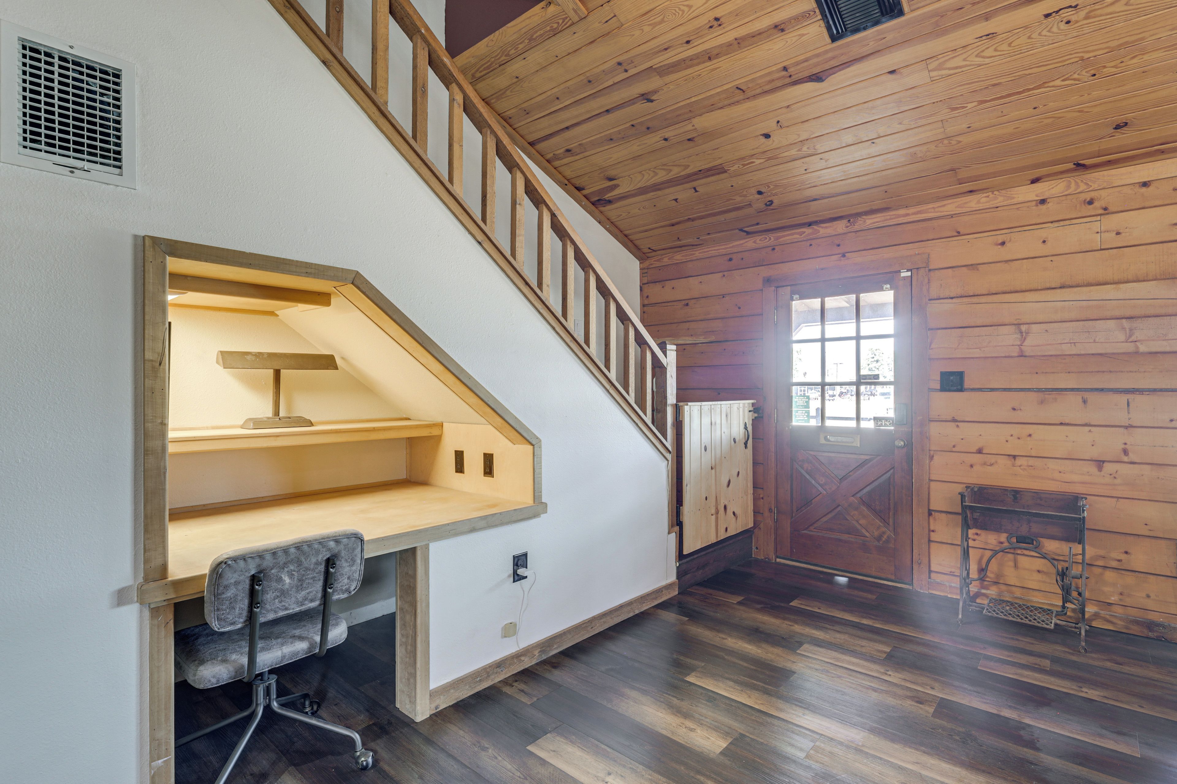 Una habitación con escalera de madera, un escritorio con silla y una puerta con ventana.
