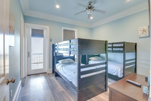 Une chambre avec des lits superposés, un bureau en bois, un ventilateur de plafond et une porte donnant sur un balcon.