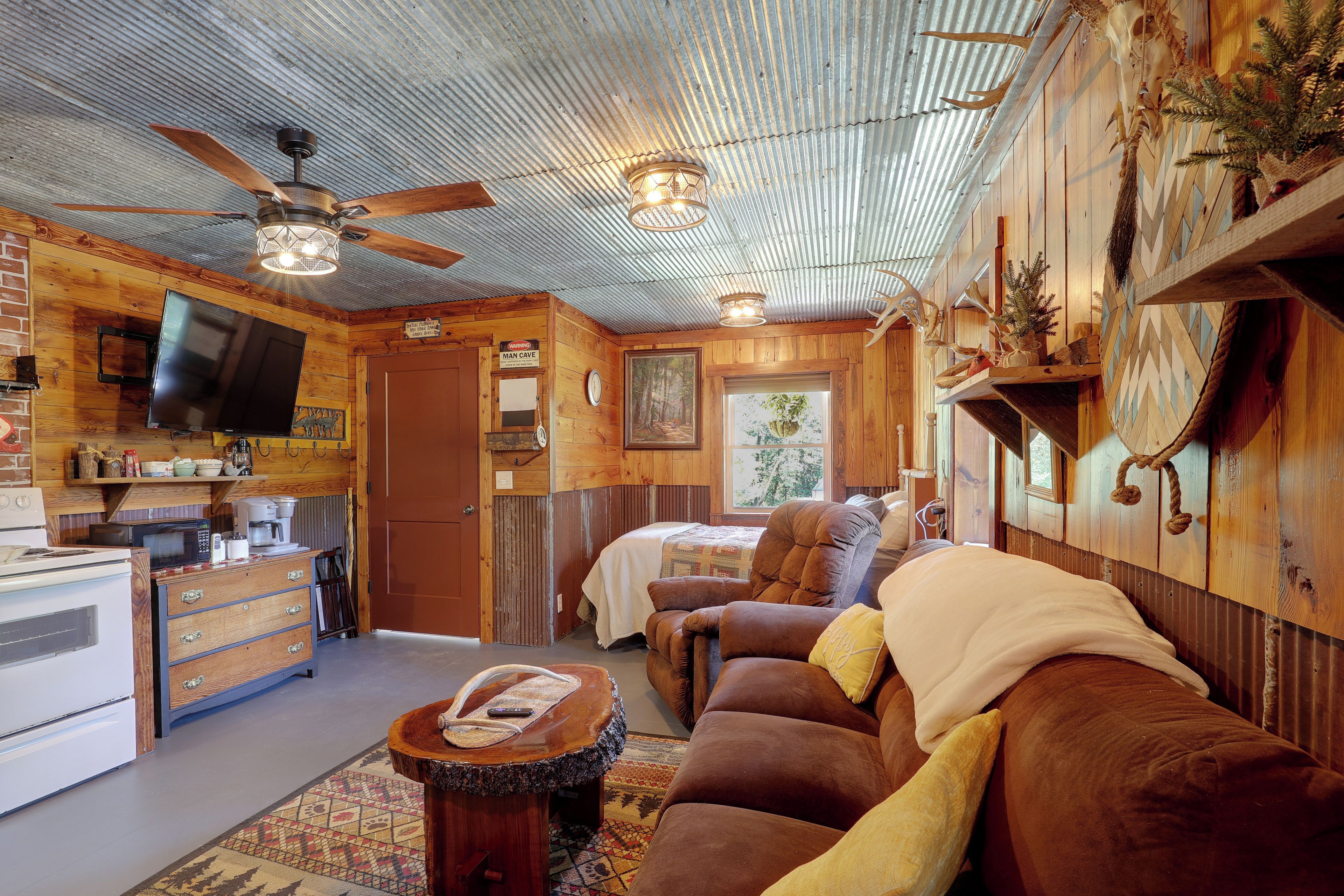 A cozy cabin interior with a wooden interior, a ceiling fan, a flat-screen TV, a kitchenette, a sofa, and a bed.