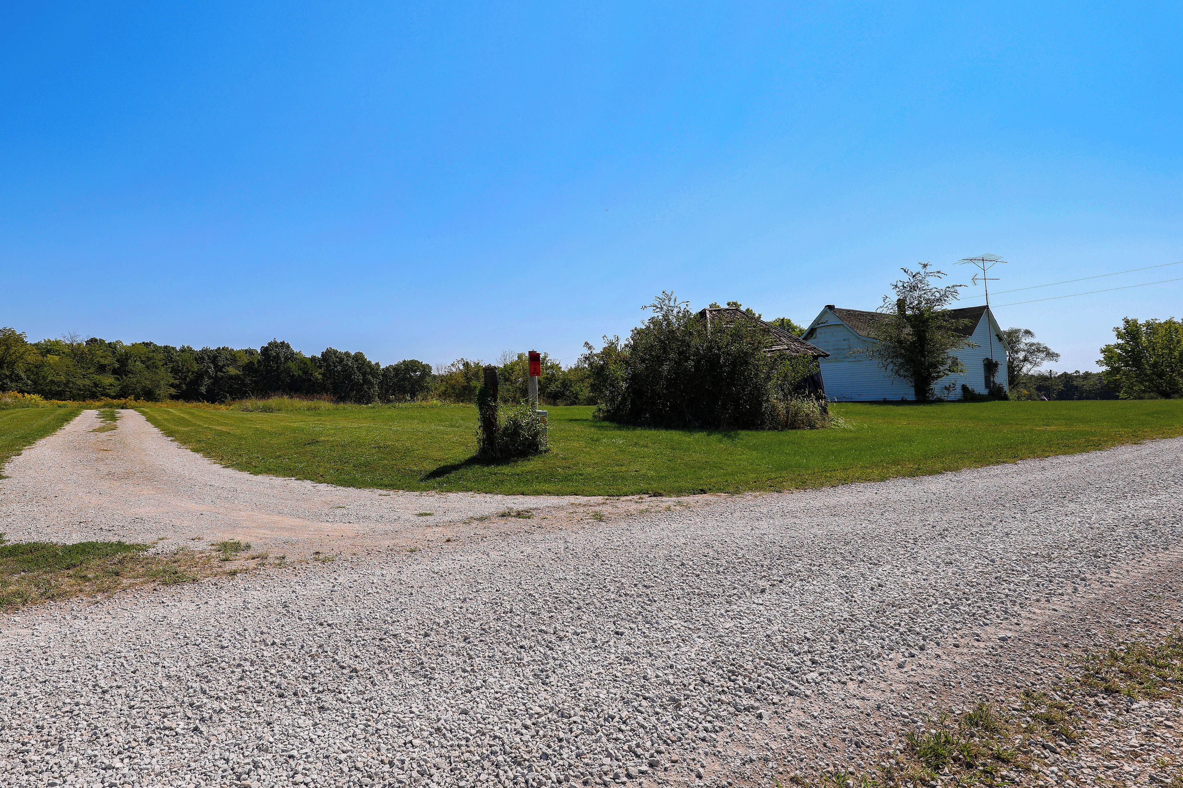 A gravel road leading to a white house with a chimney, surrounded by greenery and trees.