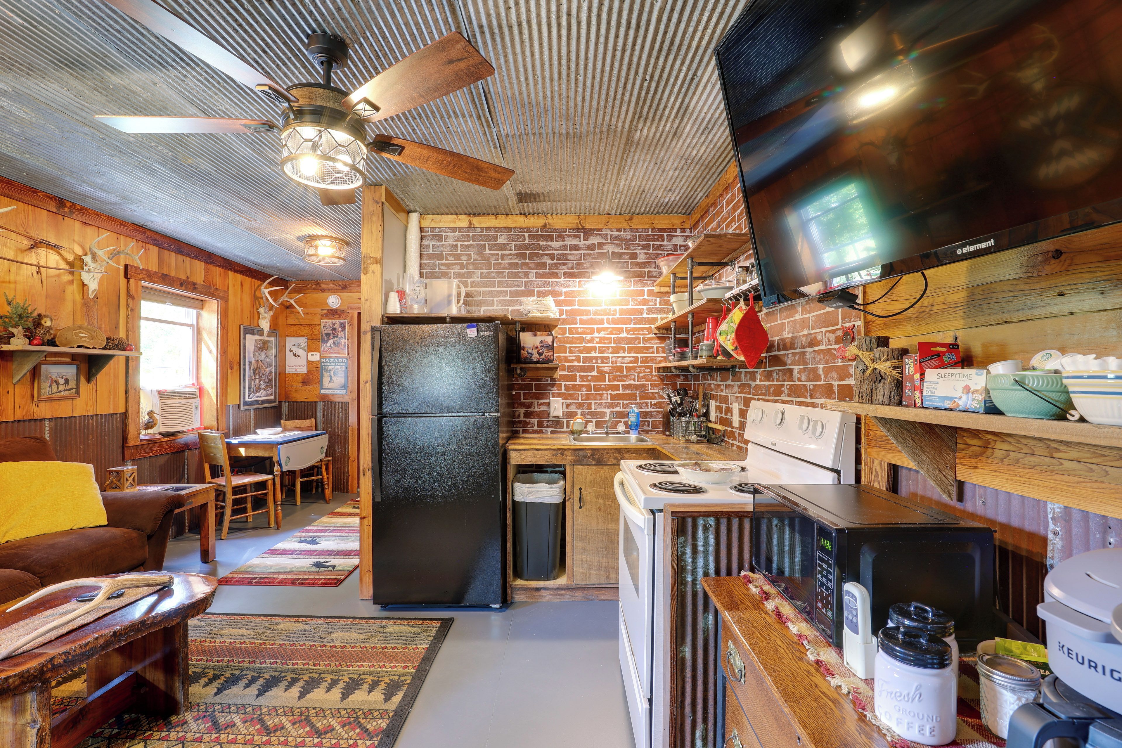 A rustic kitchen with a brick wall, wooden cabinets, a refrigerator, a stove, and a television mounted above the counter.