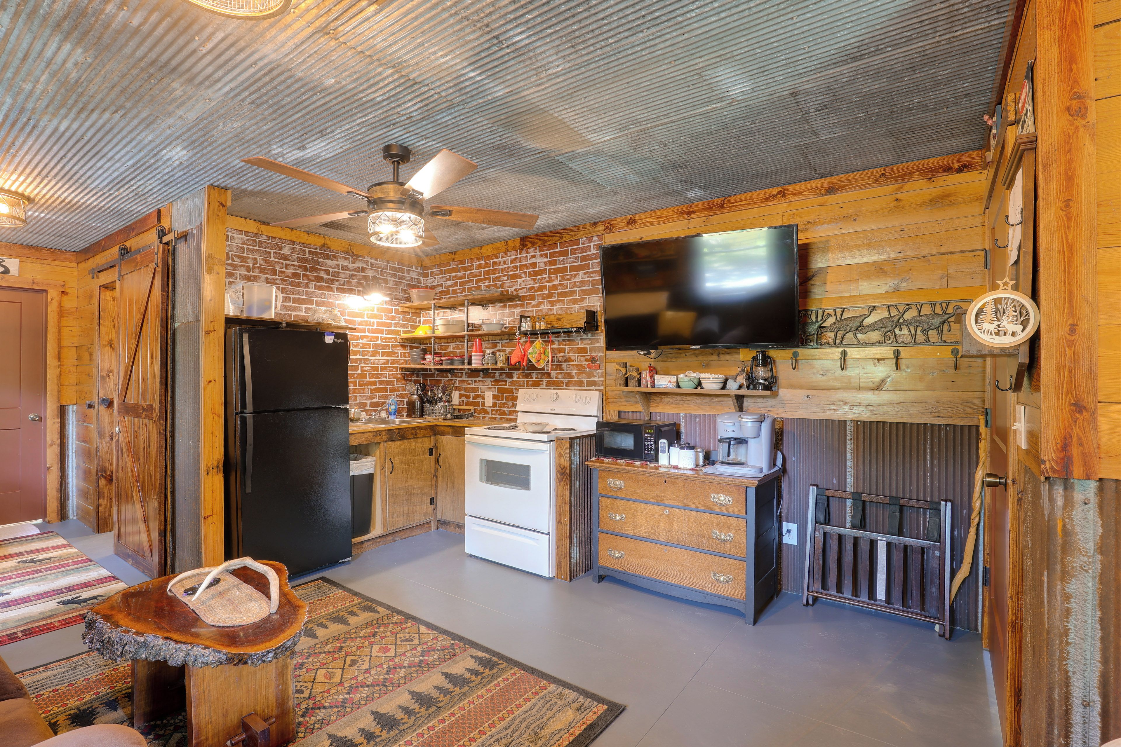 A rustic kitchen with a brick wall, wooden cabinets, a black refrigerator, a white stove, and a flat-screen TV mounted above the counter.