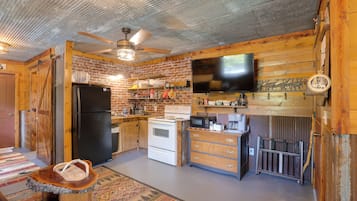 A rustic kitchen with a brick wall, wooden cabinets, a black refrigerator, a white stove, and a flat-screen TV mounted above the counter.