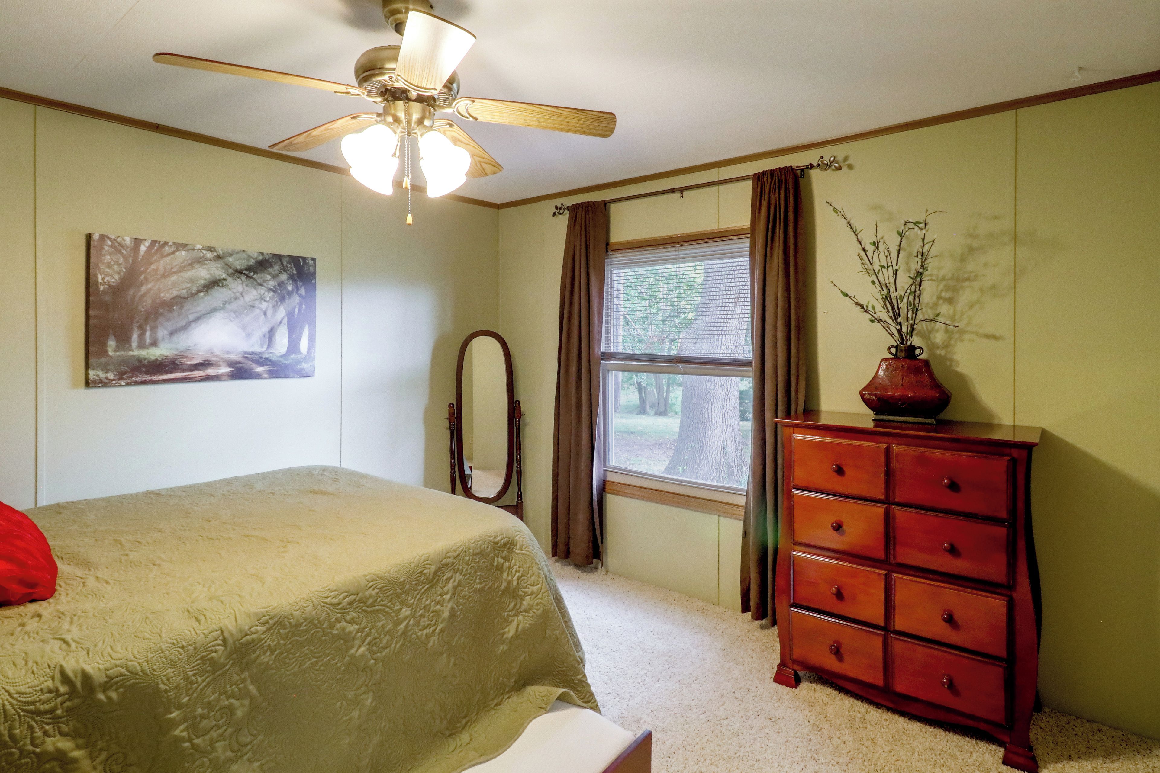 A bedroom with a bed, a dresser, a window with curtains, a ceiling fan, and a painting on the wall.