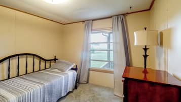 Une petite chambre avec un lit simple, une tête de lit en bois, une table de chevet avec une lampe, une fenêtre avec des rideaux et une vue sur un espace verdoyant.