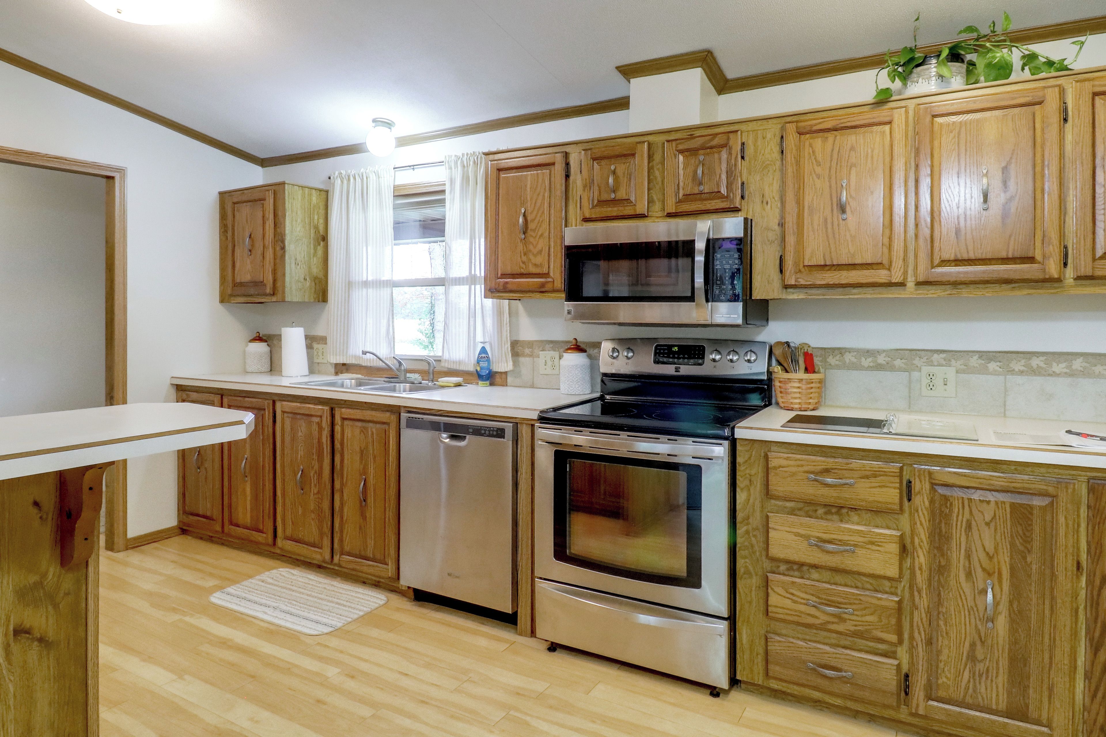 A kitchen with wooden cabinets, stainless steel appliances, a microwave, oven, dishwasher, and a window with curtains.