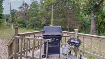 A deck with a barbecue grill and a gas tank, surrounded by a wooden railing and trees.