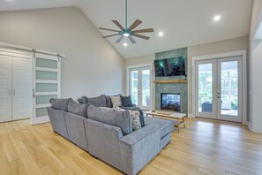 A spacious living room with a grey sectional sofa, wooden flooring, a ceiling fan, and a fireplace.
