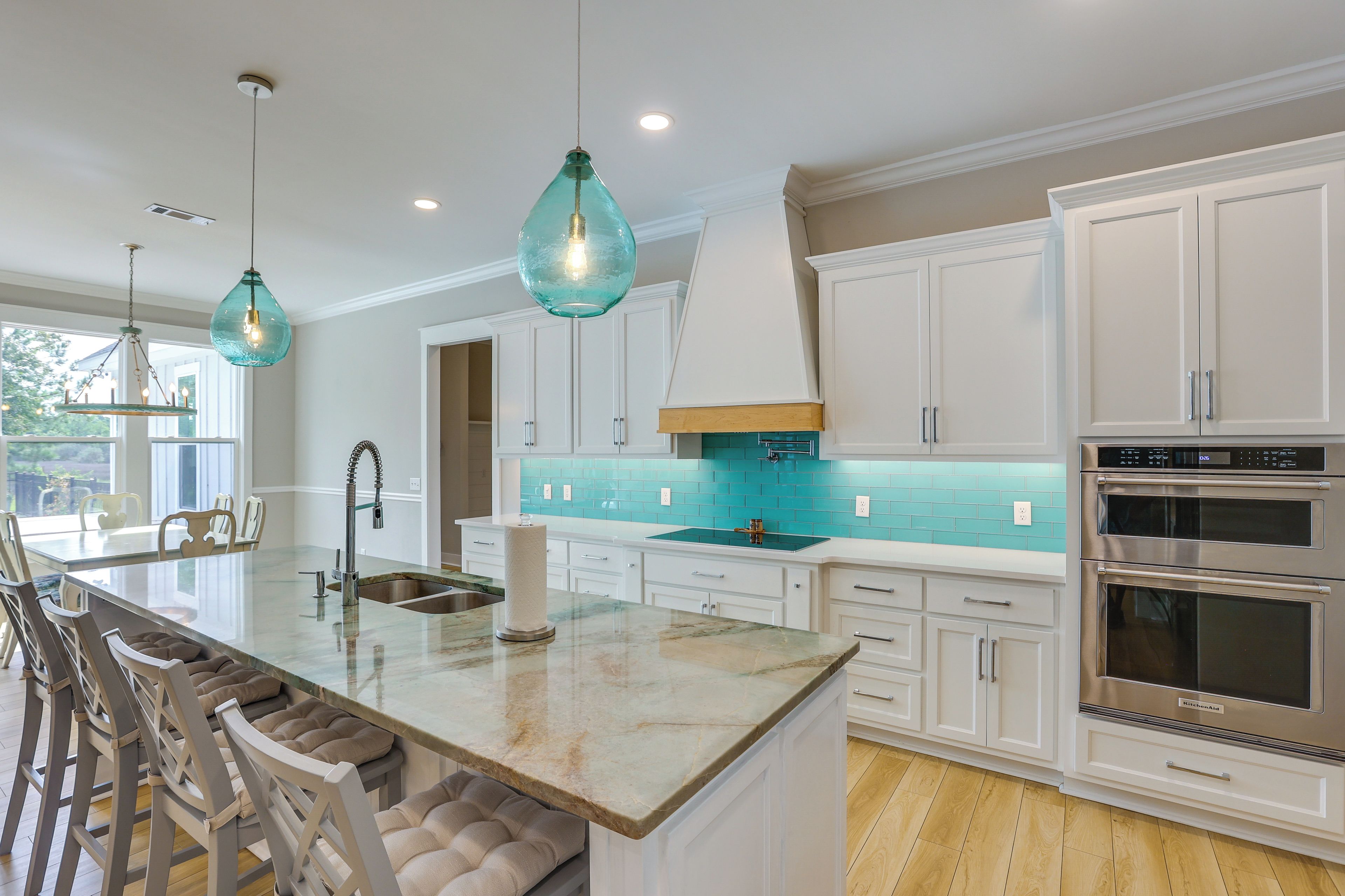 A modern kitchen with white cabinetry, a central island with stools, a stainless steel oven, and teal backsplash.