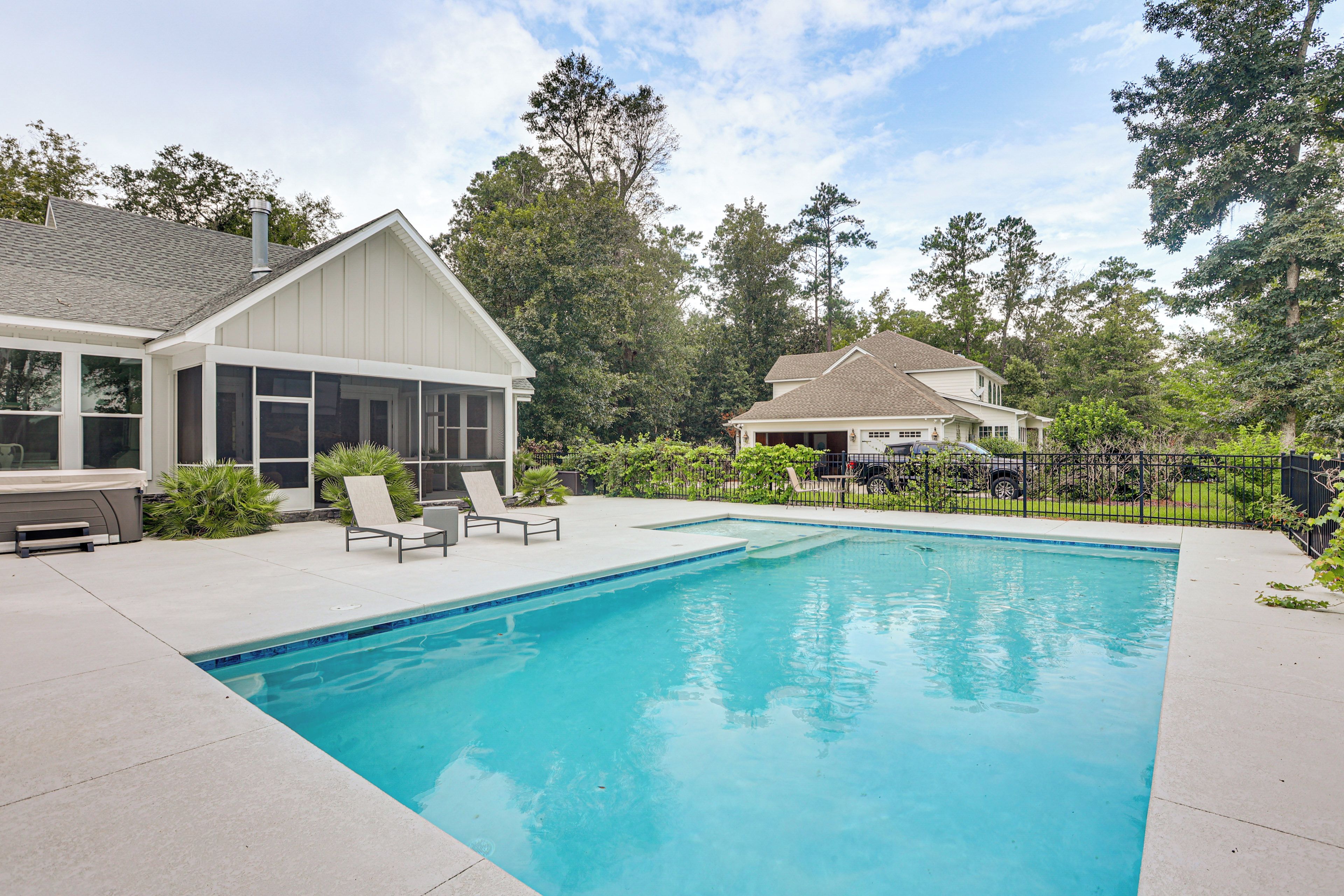 A residential swimming pool with a covered patio area, lounge chairs, and a fenced yard with trees.