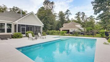 A residential swimming pool with a covered patio area, lounge chairs, and a fenced yard with trees.