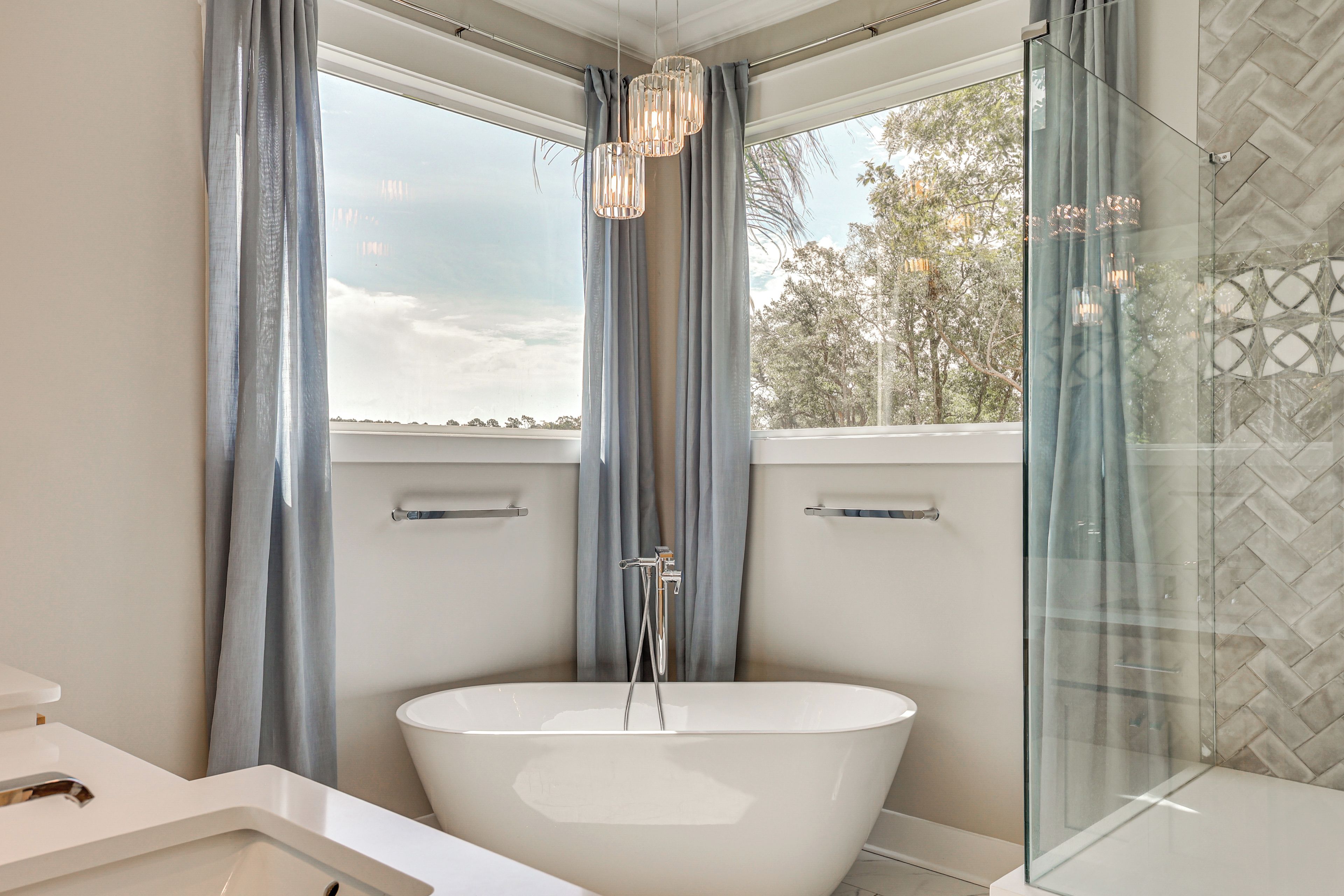 A modern bathroom with a freestanding bathtub, a glass shower enclosure, and a window with curtains.