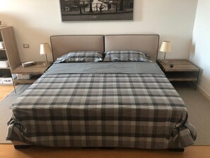 A bed with a checkered bedspread, two bedside tables with lamps, a nightstand with a remote control, and a framed picture on the wall. - Holiday Home 'Aurora Elegant Lake View' with Lake View, Shared Pool and Wi-Fi (Toscolano Maderno)