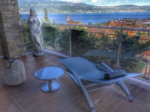 A balcony with a view of a coastal town, a statue, a lounge chair, and a small table. - Holiday Home 'Aurora Elegant Lake View' with Lake View, Shared Pool and Wi-Fi (Toscolano Maderno)