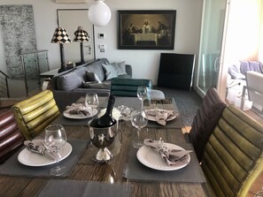 A dining area with a table set for four, a bottle of wine, and glasses. There is a sofa and a green ottoman in the background. - Holiday Home 'Aurora Elegant Lake View' with Lake View, Shared Pool and Wi-Fi (Toscolano Maderno)
