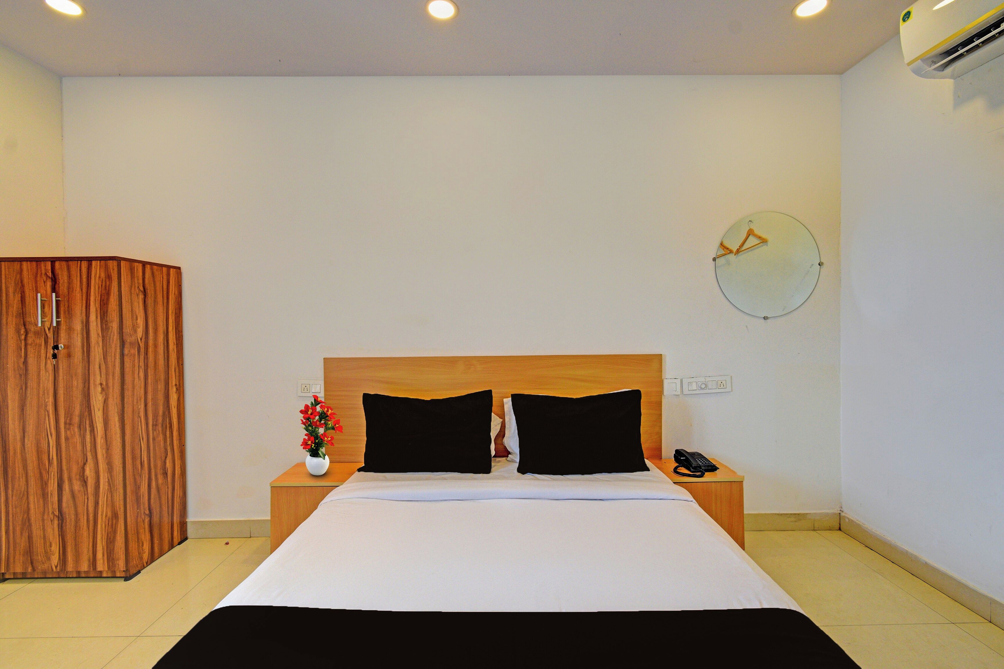 A hotel room with a bed, bedside table, a clock on the wall, and an air conditioning unit.