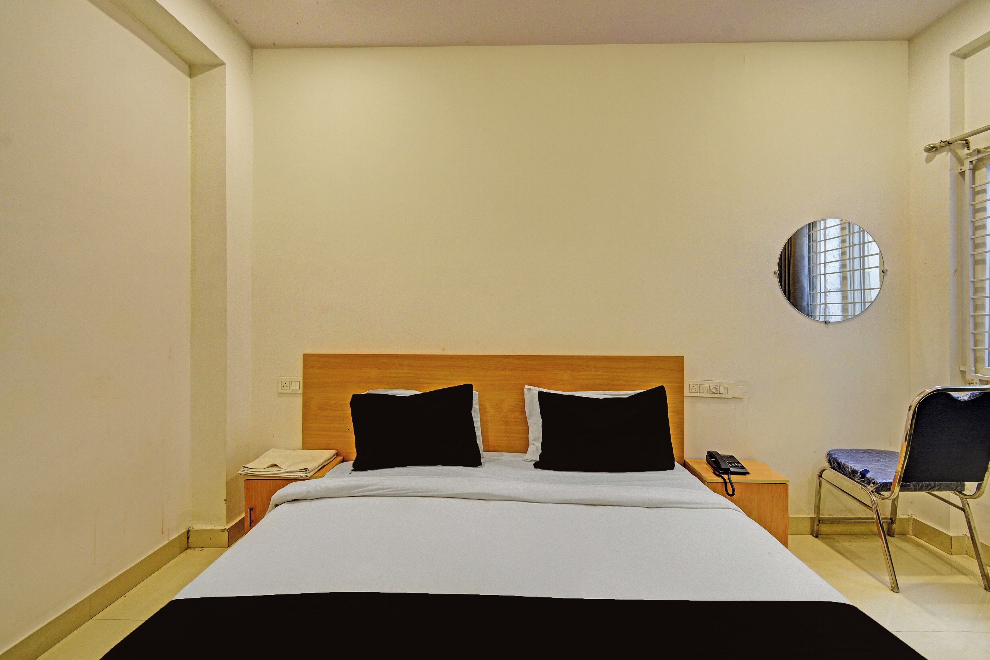 Quarto de hotel com cama, espelho redondo na parede, mesa de cabeceira com telefone e uma cadeira.
